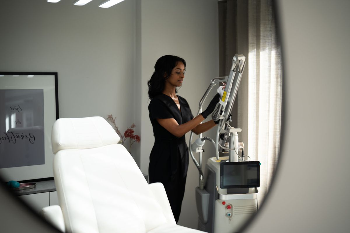 Klinikutrymme med laserapparat och behandlare i svart arbetsdräkt intill ett fönster