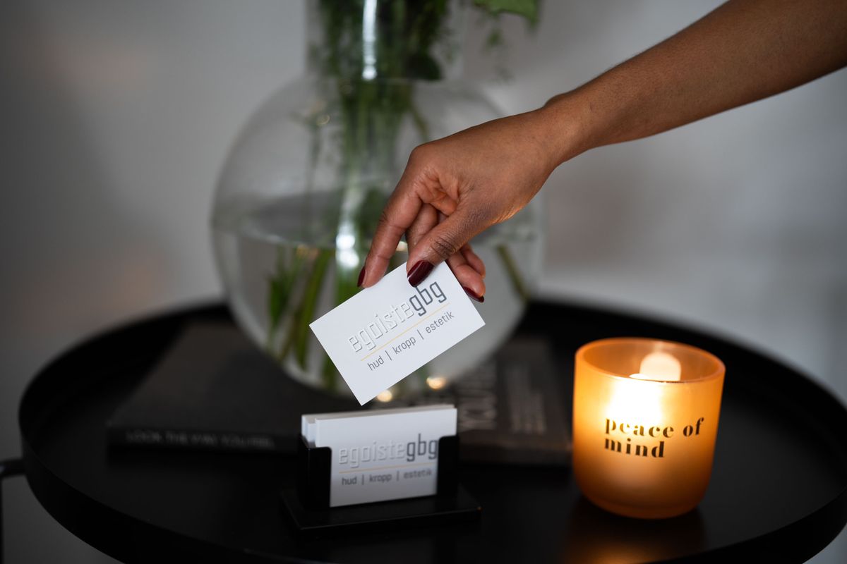 Hand som håller upp ett vitt visitkort med egoiste|gbg framför en korthållare, en glasvas med växter och ett tänt ljus